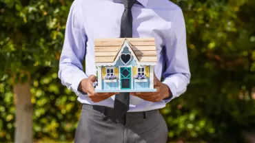 Persona sosteniendo una casa en miniatura con ventanas amarillas y flores.