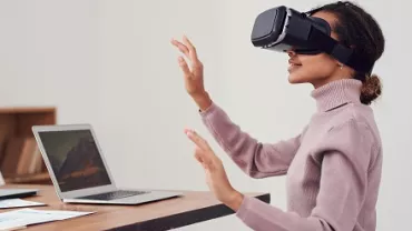 Persona usando una realidad virtual mientras está sentada frente a una computadora portátil.