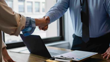 Personas dándose la mano en una oficina junto a un portátil sobre la mesa.