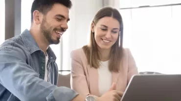 Dos personas están sentadas en una mesa frente a un portátil, sonriendo. 