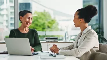 Dos mujeres conversando en una mesa, con un portátil abierto entre ellas.