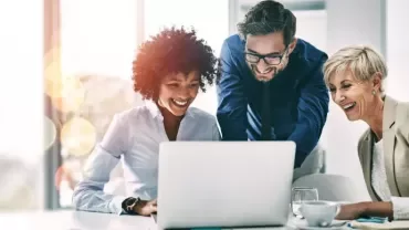 dos mujeres y un hombre mirando a un ordenador mientras sonrien