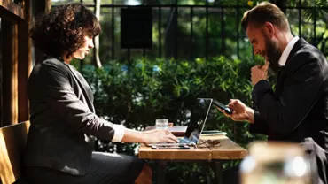 Dos personas trabajando en un café al aire libre con computadoras portátiles.