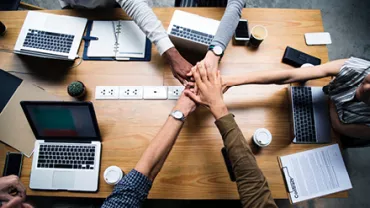 Personas uniendo sus manos sobre una mesa de trabajo con laptops y café.