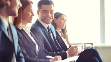 Personas sentadas en traje, sonriendo y escribiendo en un entorno corporativo.