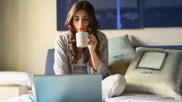 Persona tomando café frente a una laptop en la cama.