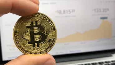 Moneda de Bitcoin frente a laptop con gráfico en pantalla.