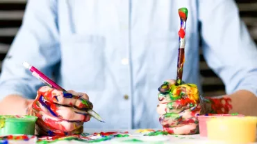 Manos cubiertas de pintura sosteniendo un pincel y un bolígrafo.