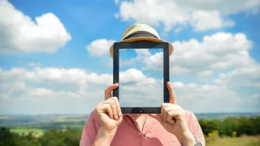 Persona sosteniendo una tablet con imagen de nubes frente a su cara, en un campo.