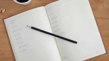 Un cuaderno abierto con un lápiz negro y una taza de café en una mesa de madera.