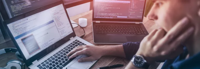 Persona escribiendo código en una computadora portátil, dos pantallas adicionales en el fondo.