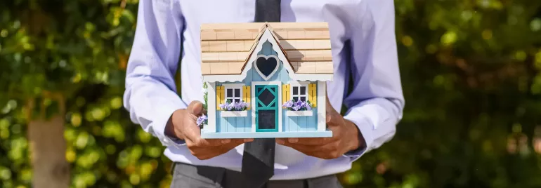 Persona sosteniendo una casa en miniatura con ventanas amarillas y flores.