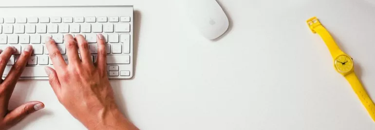 Manos escribiendo en un teclado junto a un reloj amarillo.