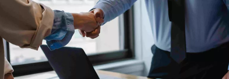 Personas dándose la mano en una oficina junto a un portátil sobre la mesa.