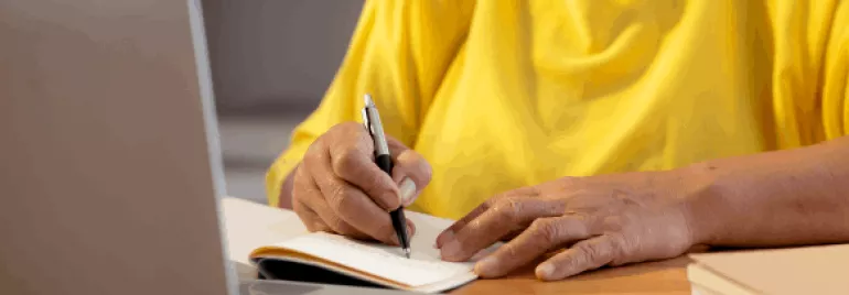 Persona escribiendo en un cuaderno junto a una laptop.