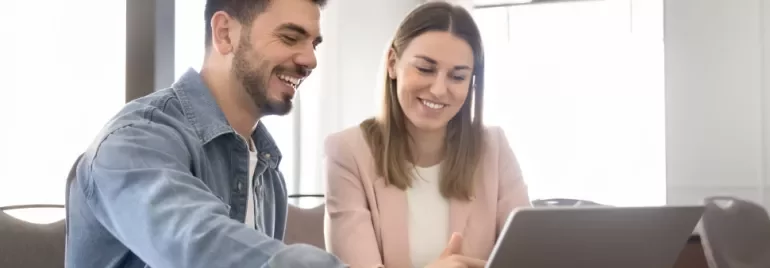 Dos personas están sentadas en una mesa frente a un portátil, sonriendo. 