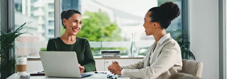 Dos mujeres conversando en una mesa, con un portátil abierto entre ellas.