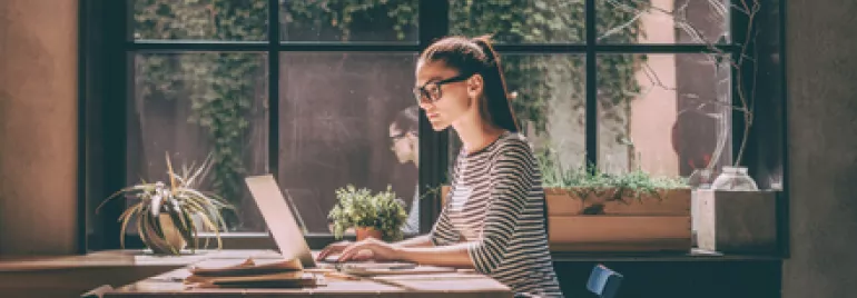 Persona con gafas trabajando en una computadora portátil junto a una ventana.