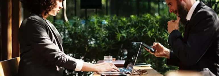 Dos personas trabajando en un café al aire libre con computadoras portátiles.