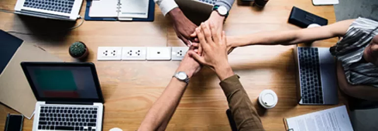 Personas uniendo sus manos sobre una mesa de trabajo con laptops y café.