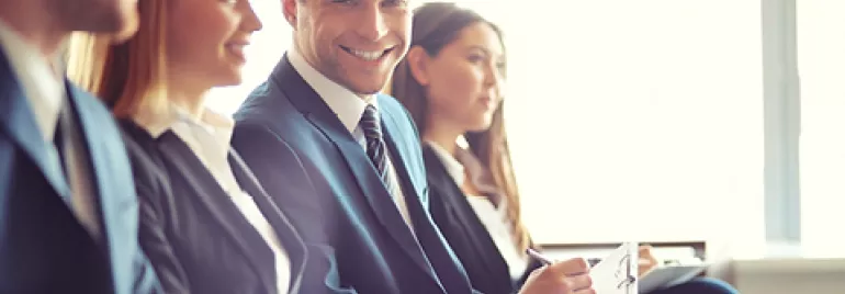 Personas sentadas en traje, sonriendo y escribiendo en un entorno corporativo.