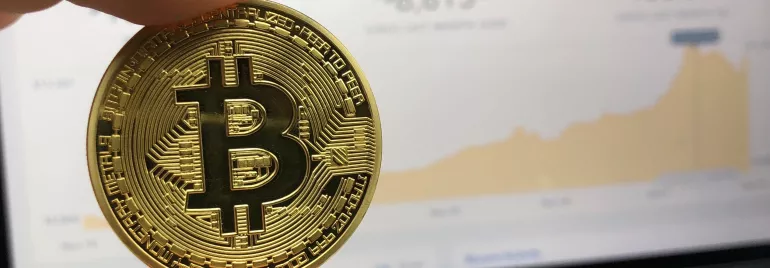 Moneda de Bitcoin frente a laptop con gráfico en pantalla.