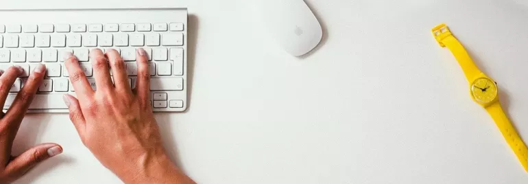 Manos escribiendo en teclado blanco sobre escritorio blanco con reloj amarillo.