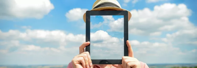 Persona sosteniendo una tablet con imagen de nubes frente a su cara, en un campo.