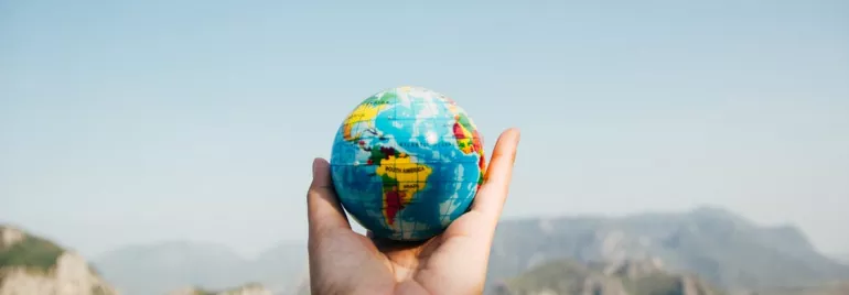 Una mano sostiene un pequeño globo terráqueo frente a un paisaje montañoso.