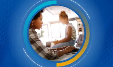 Persona trabajando en una computadora mientras otra persona está sentada cerca.