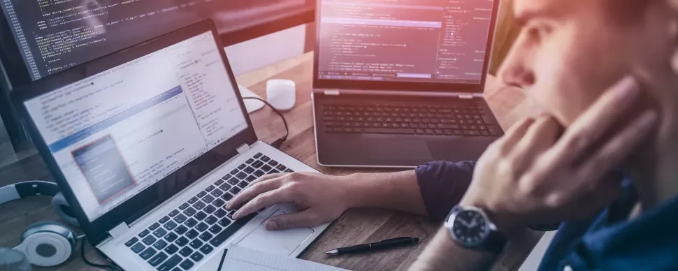 Persona escribiendo código en una computadora portátil, dos pantallas adicionales en el fondo.