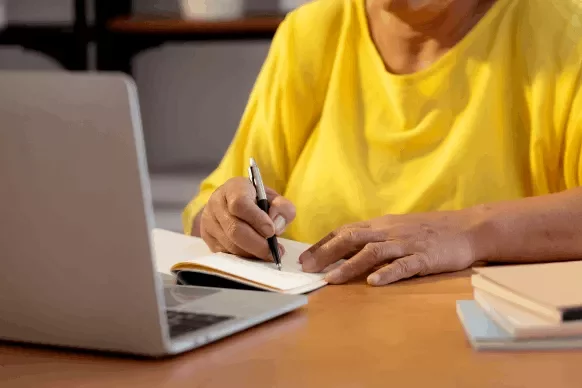 Persona escribiendo en un cuaderno junto a una laptop.