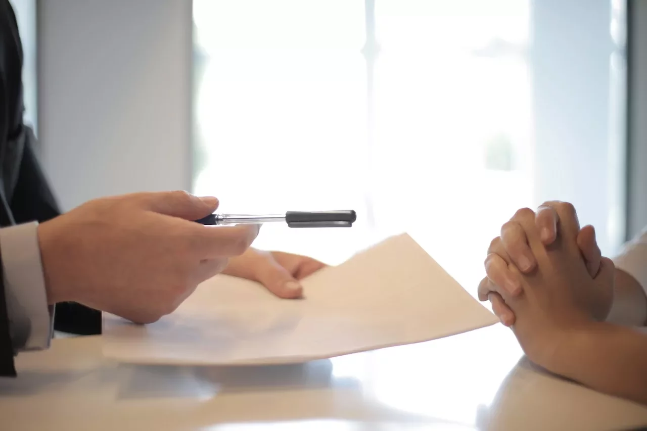 Mano entregando un bolígrafo y documento en un escritorio.