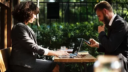 Dos personas trabajando en un café al aire libre con computadoras portátiles.