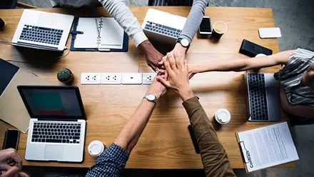 Personas uniendo sus manos sobre una mesa de trabajo con laptops y café.