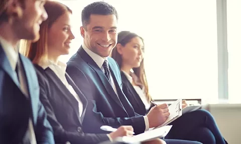 Personas sentadas en traje, sonriendo y escribiendo en un entorno corporativo.