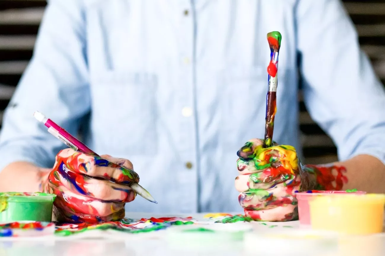 Manos cubiertas de pintura sosteniendo un pincel y un bolígrafo.