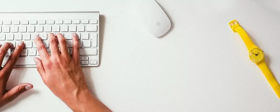 Manos escribiendo en teclado blanco sobre escritorio blanco con reloj amarillo.