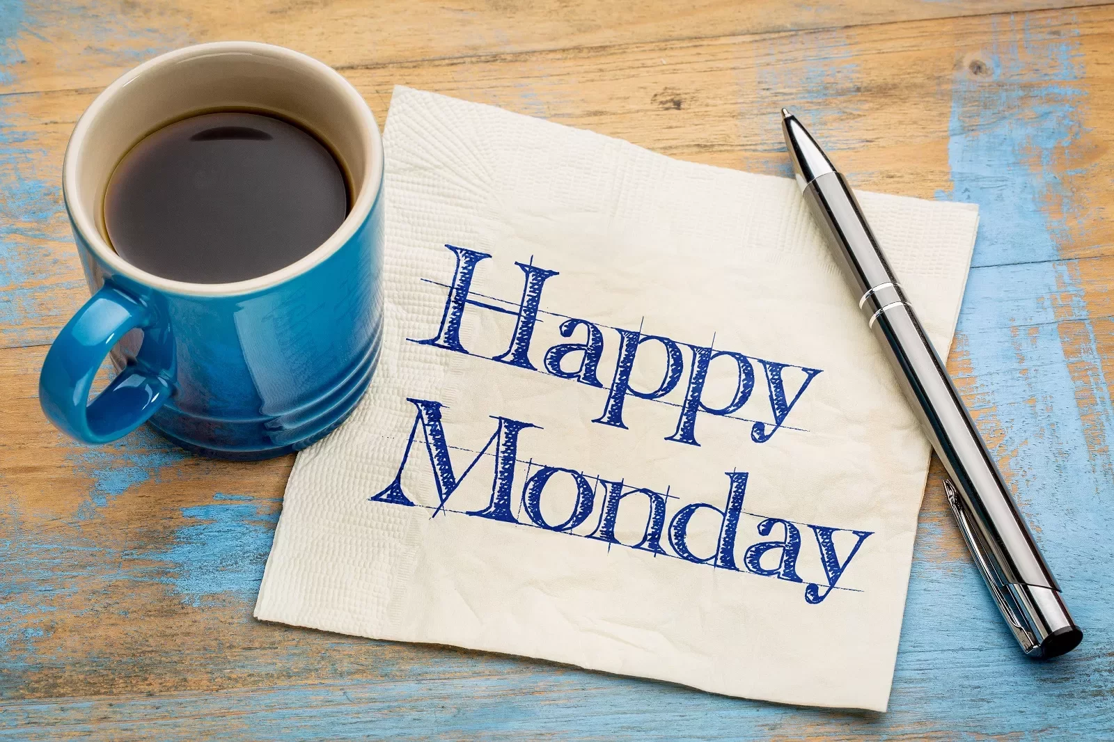 Taza de café y bolígrafo al lado de servilleta que dice 'Happy Monday'.