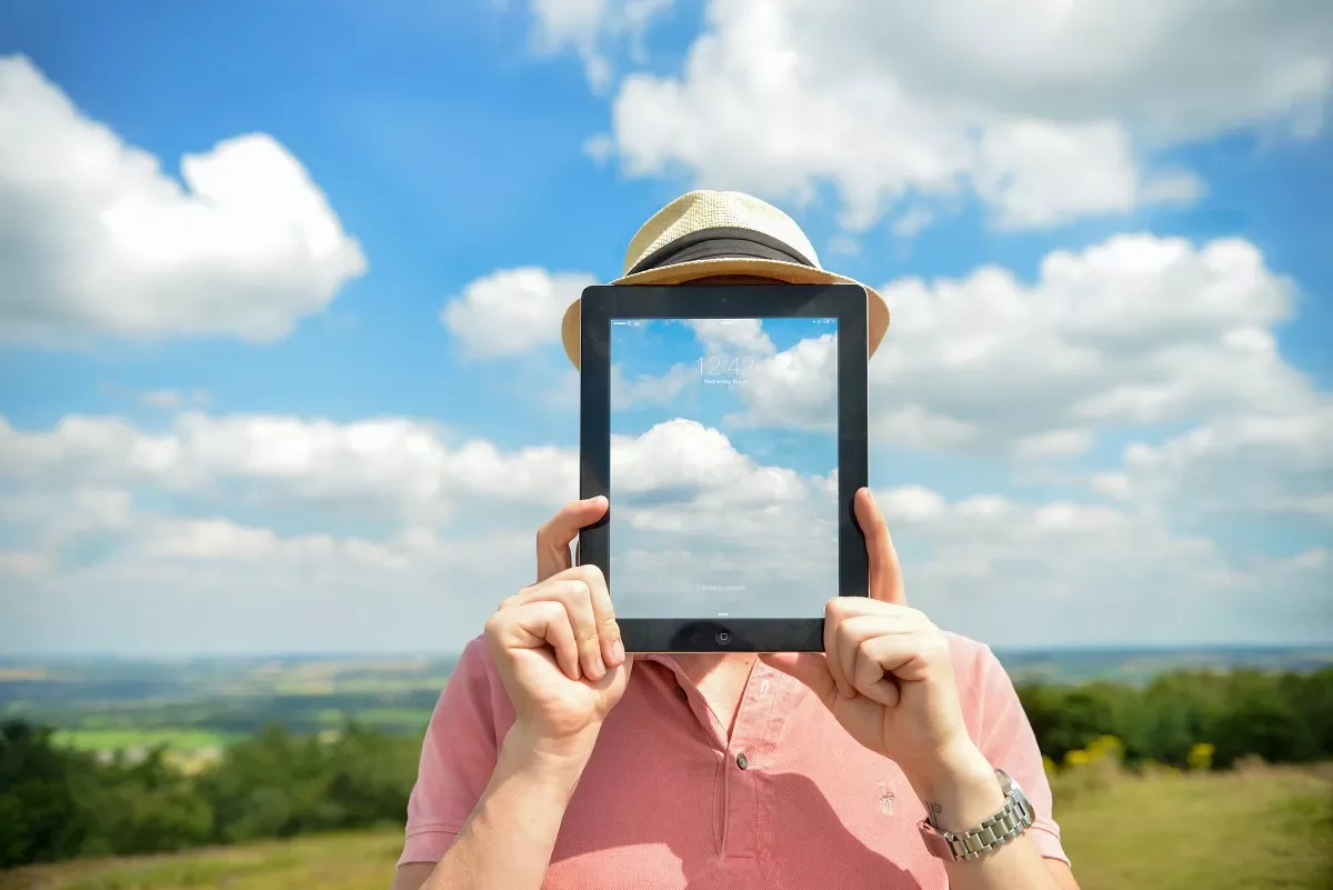 Persona sosteniendo una tablet con imagen de nubes frente a su cara, en un campo.